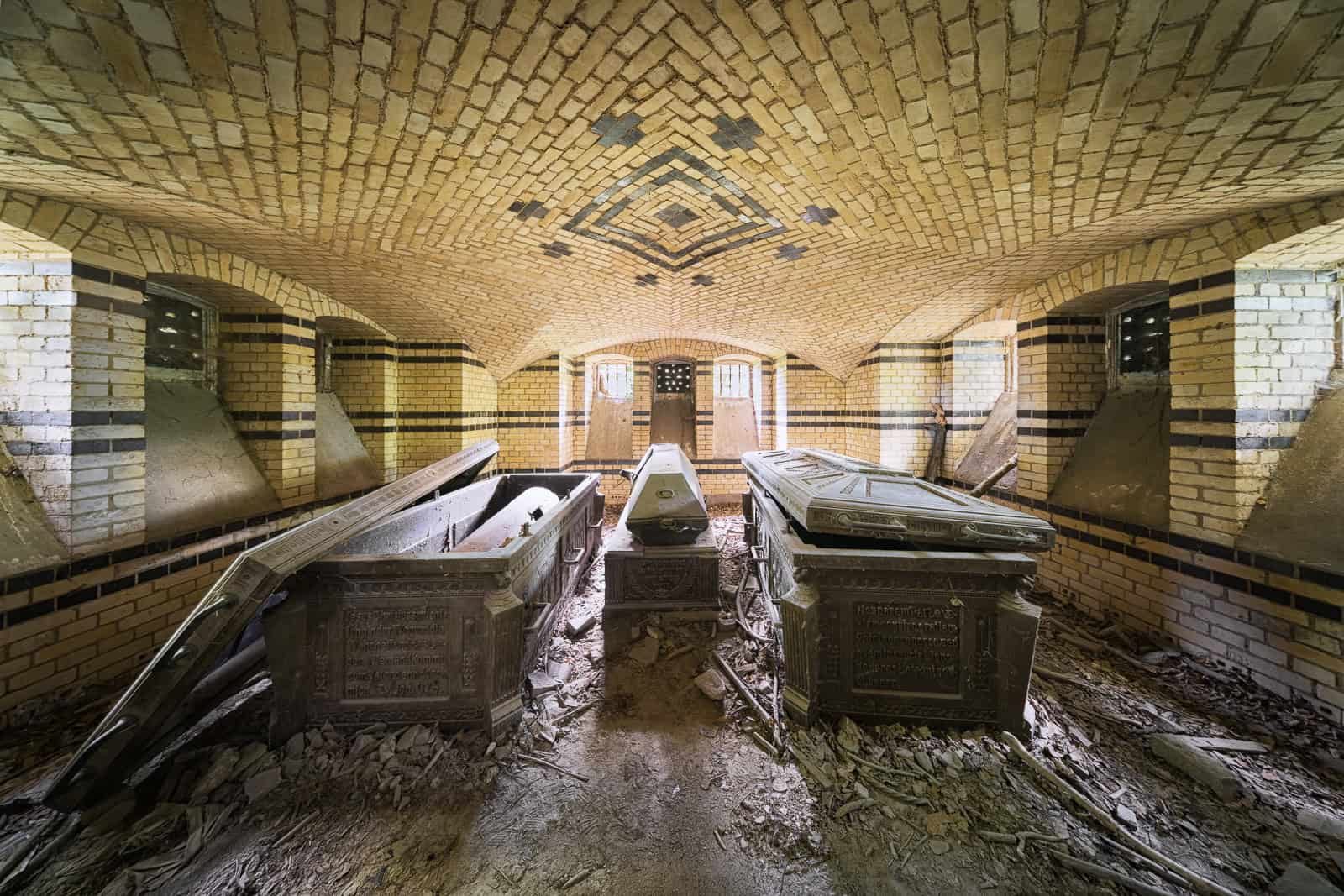 An abandoned mausoleum with overturned coffins and debris scattered across the floor under a vaulted brick ceiling.